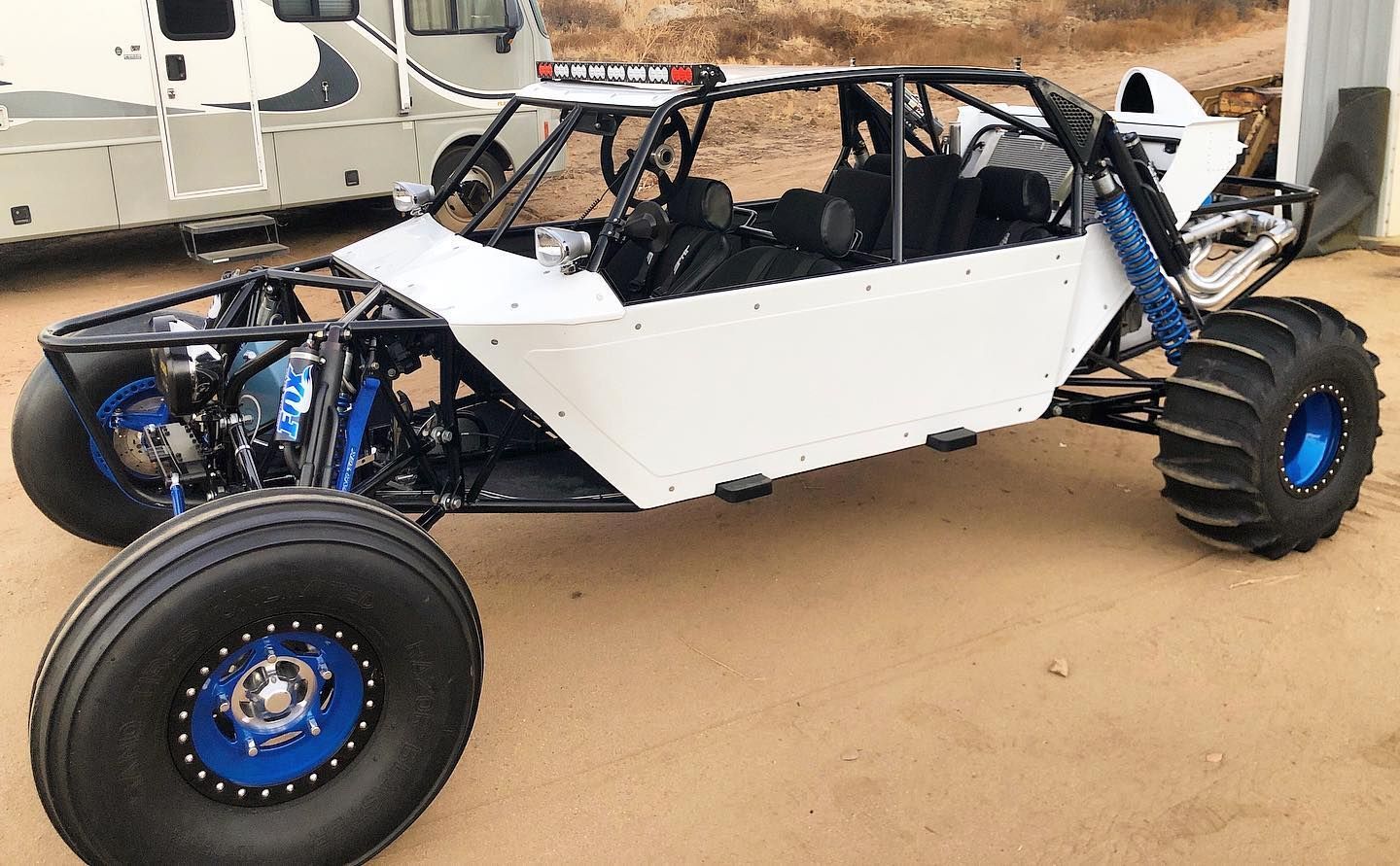 A white four-seat sand rail dune buggy with blue wheels and large off-road tires parked on a dirt lot.