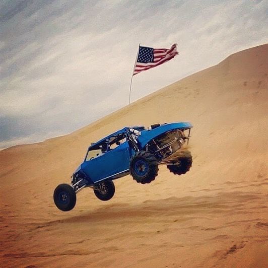 A blue sand rail jumps over a sandy dune with an American flag flying in the background.