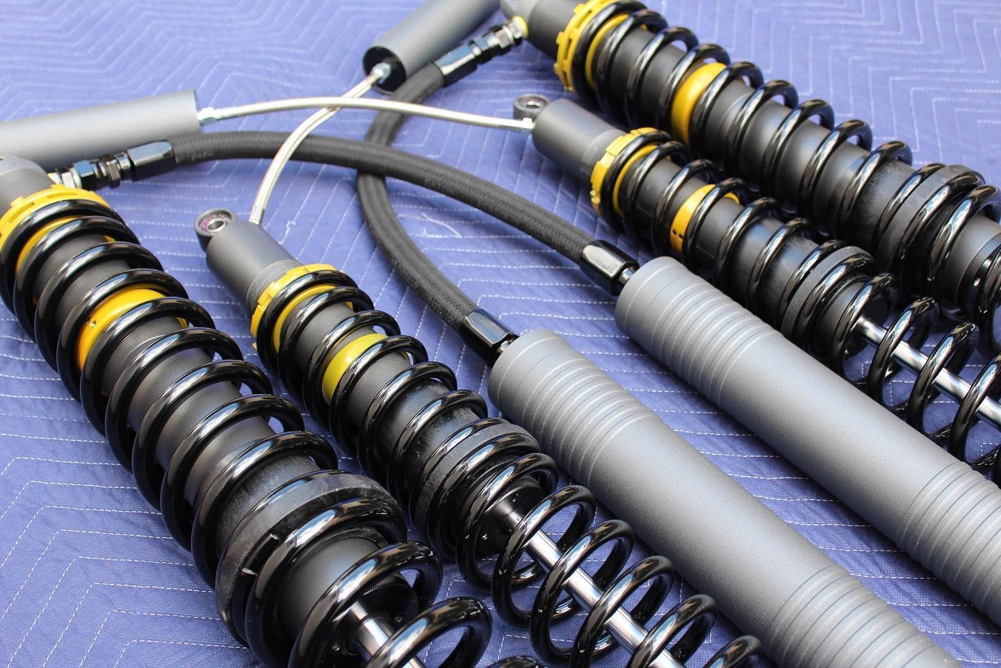 Two high-performance vehicle shock absorbers with black springs and external yellow reservoirs, set against a blue fabric.