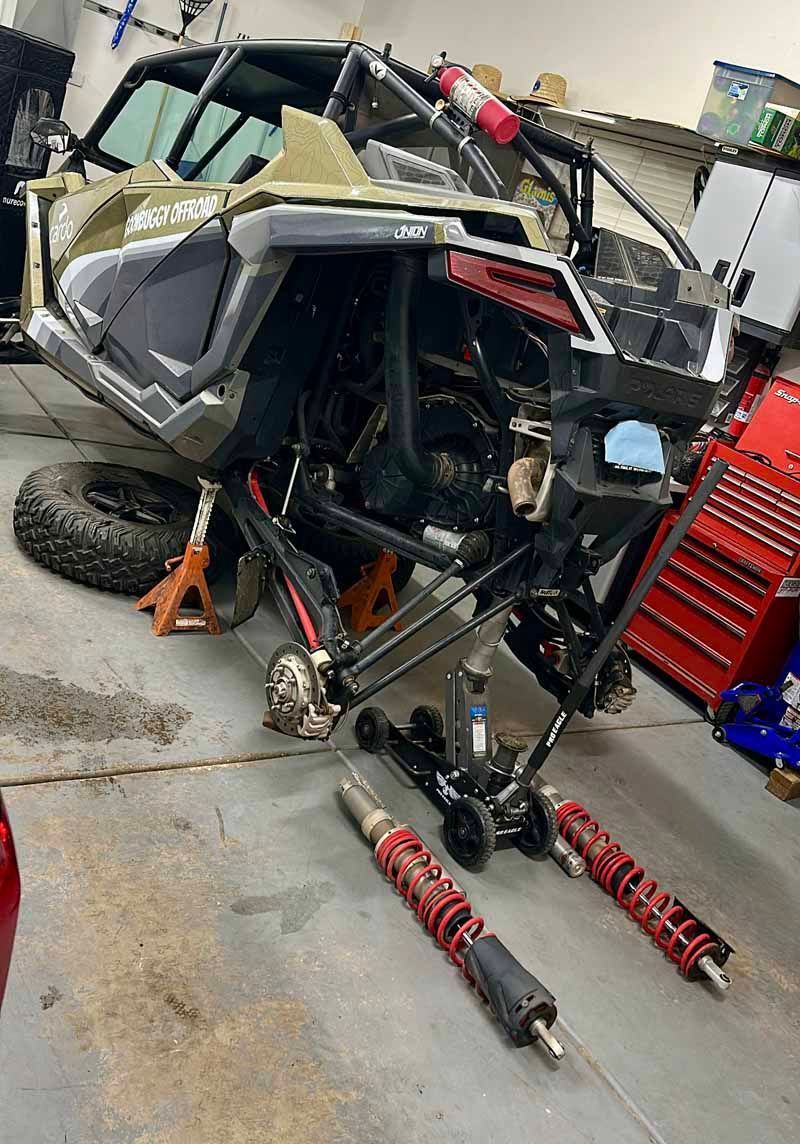 A side-by-side off-road vehicle jacked up in a garage with its rear shocks removed and lying on the floor.
