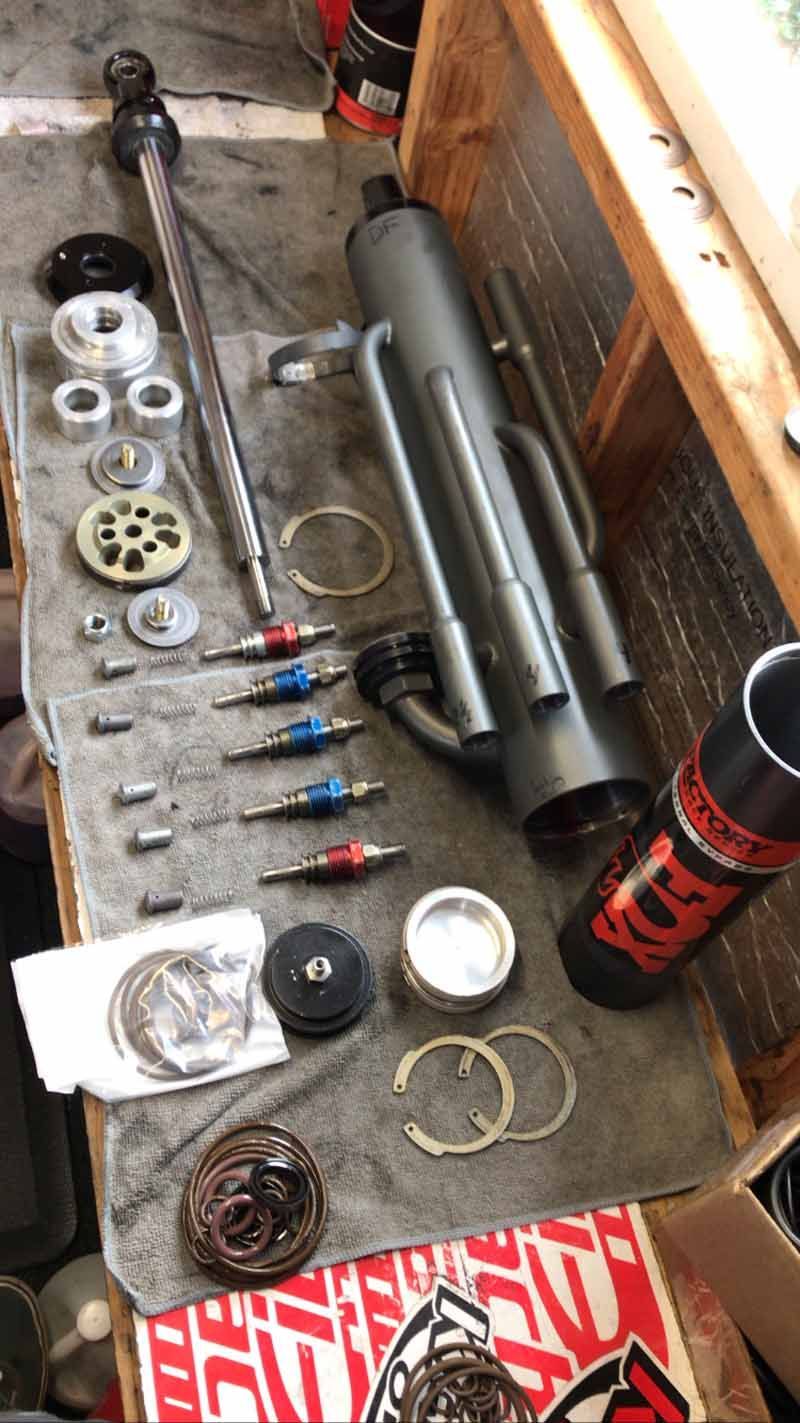 Disassembled Fox suspension shock components laid out on a workbench, including a piston rod, seals, and a housing.
