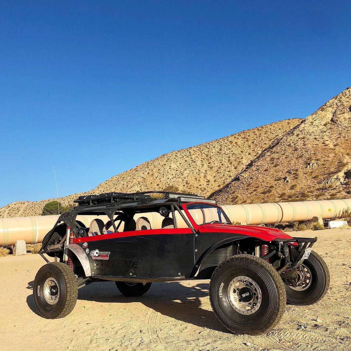 A two-tone red and black off-road buggy parked on a sandy desert landscape in front of a mountain and large pipe.
