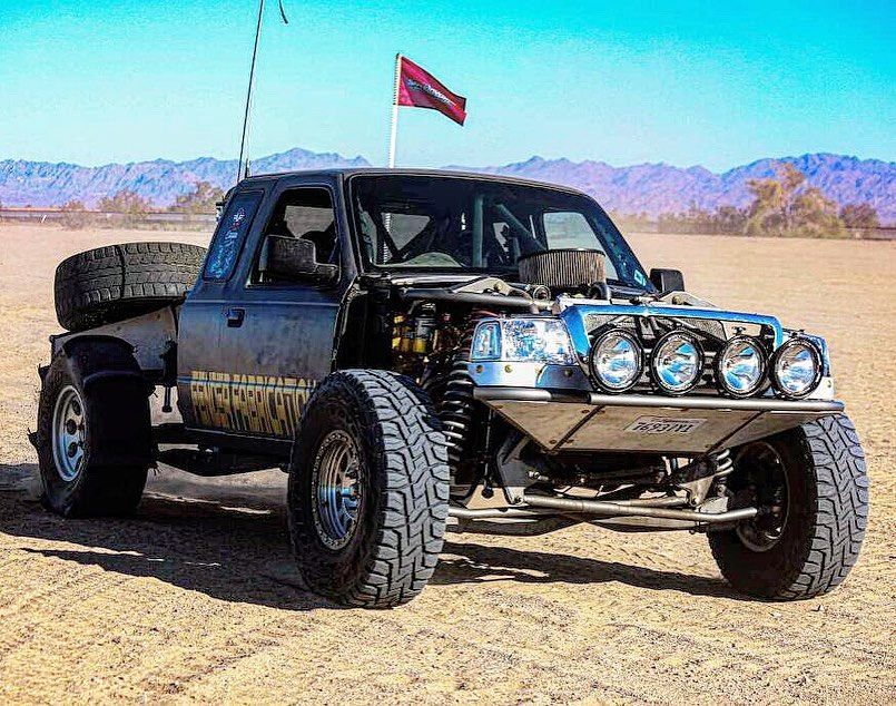 A modified, rugged off-road desert racing truck with large tires and exposed engine parts sits on a sandy desert landscape.