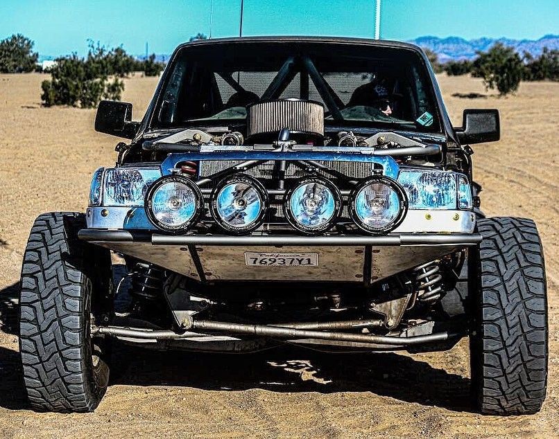 A modified off-road truck sits on a desert landscape, featuring a custom metal bumper with four large spotlights.