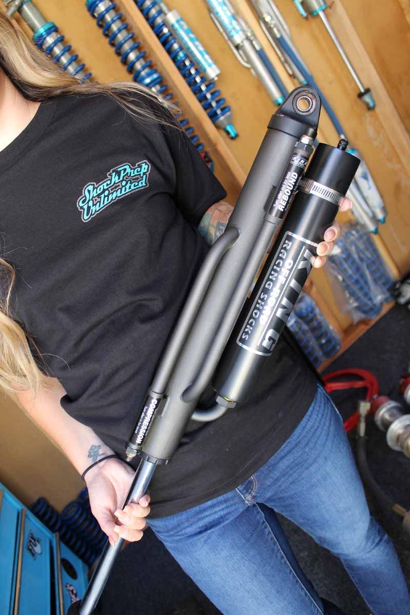 A person holds a large, black off-road shock absorber in a workshop with various automotive parts visible in the background.