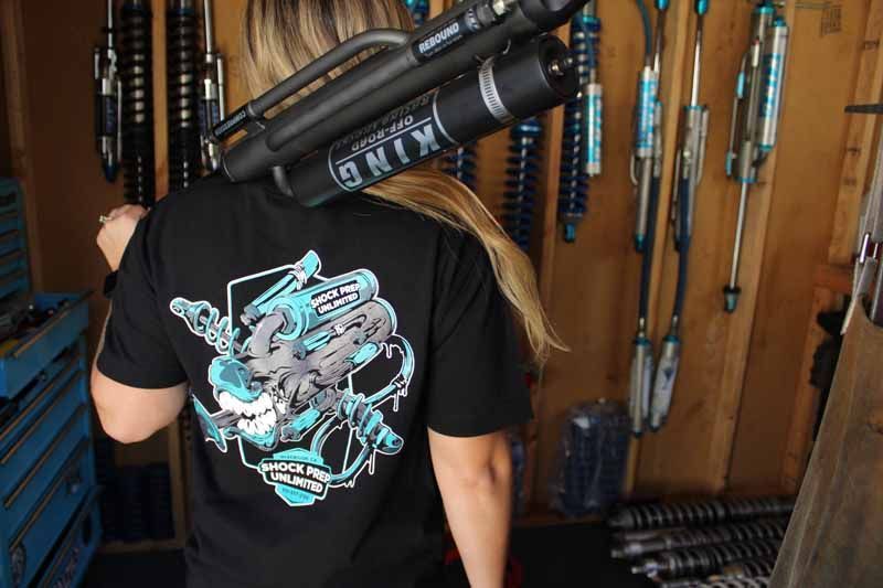 A person wears a black t-shirt with a blue graphic, carrying a large King off-road shock absorber in a workshop.