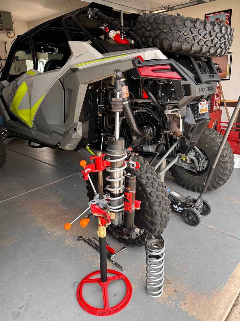 A UTV raised on a jack in a garage with one of its shock absorbers removed and placed on a spring compressor tool.