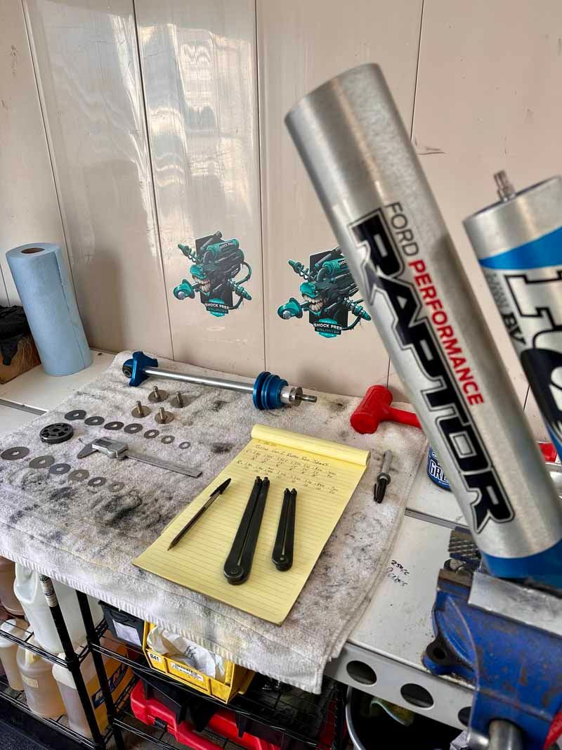 A work surface holds a Ford Performance Raptor shock, a notepad with tools, hardware, and a roll of blue shop towels.