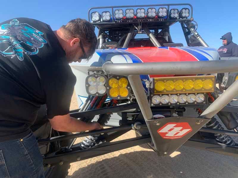 A person works on the suspension of a red, white, and blue off-road racing vehicle in a desert setting.