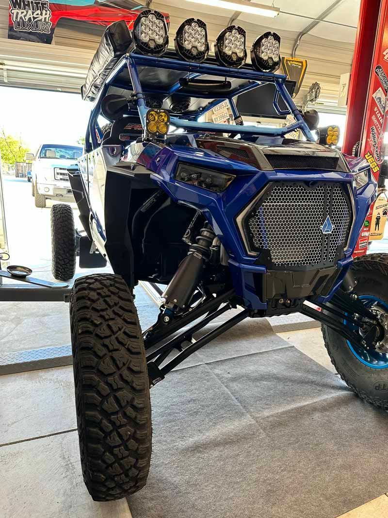 A blue Polaris RZR off-road vehicle with large tires, a roof-mounted light bar, and grille guard in a garage setting.