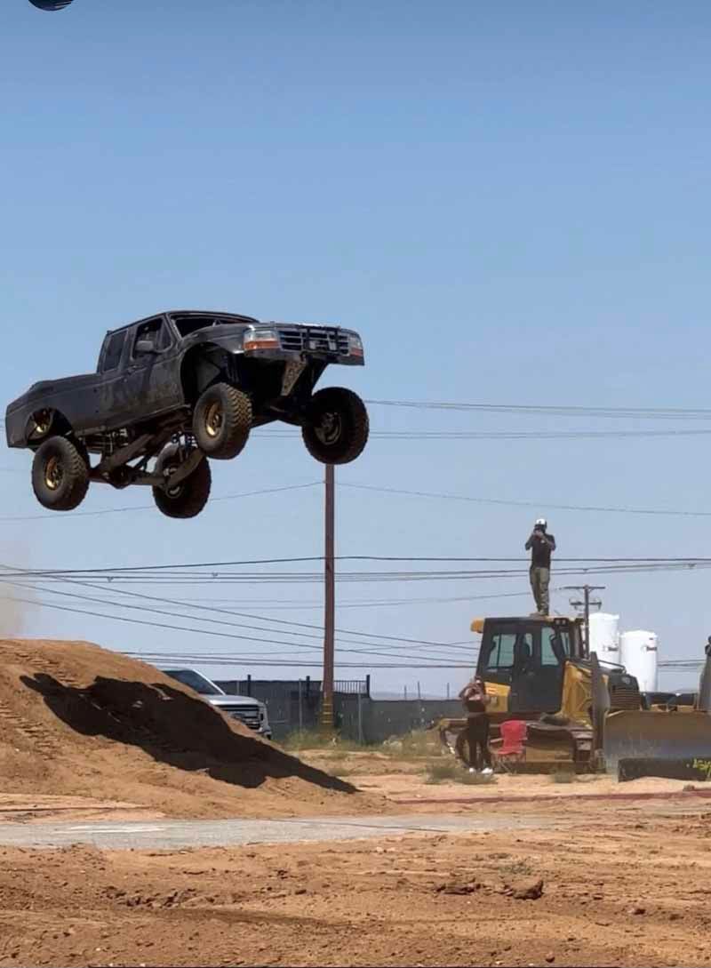 A dark-colored off-road truck jumps mid-air over a dirt mound as a person stands on a nearby bulldozer.