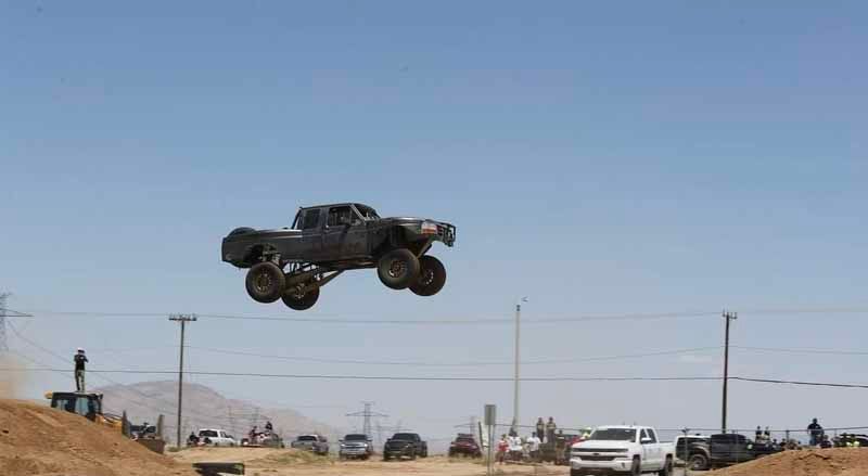 A dark off-road truck jumps high over a dirt ramp with a desert background and parked spectator vehicles.