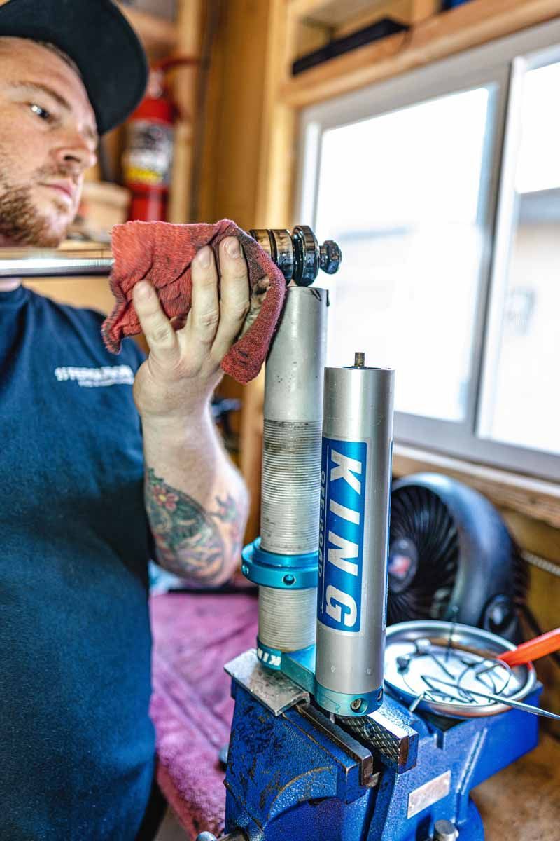 A person wipes down a King shock absorber held in a blue workbench vise inside a garage.