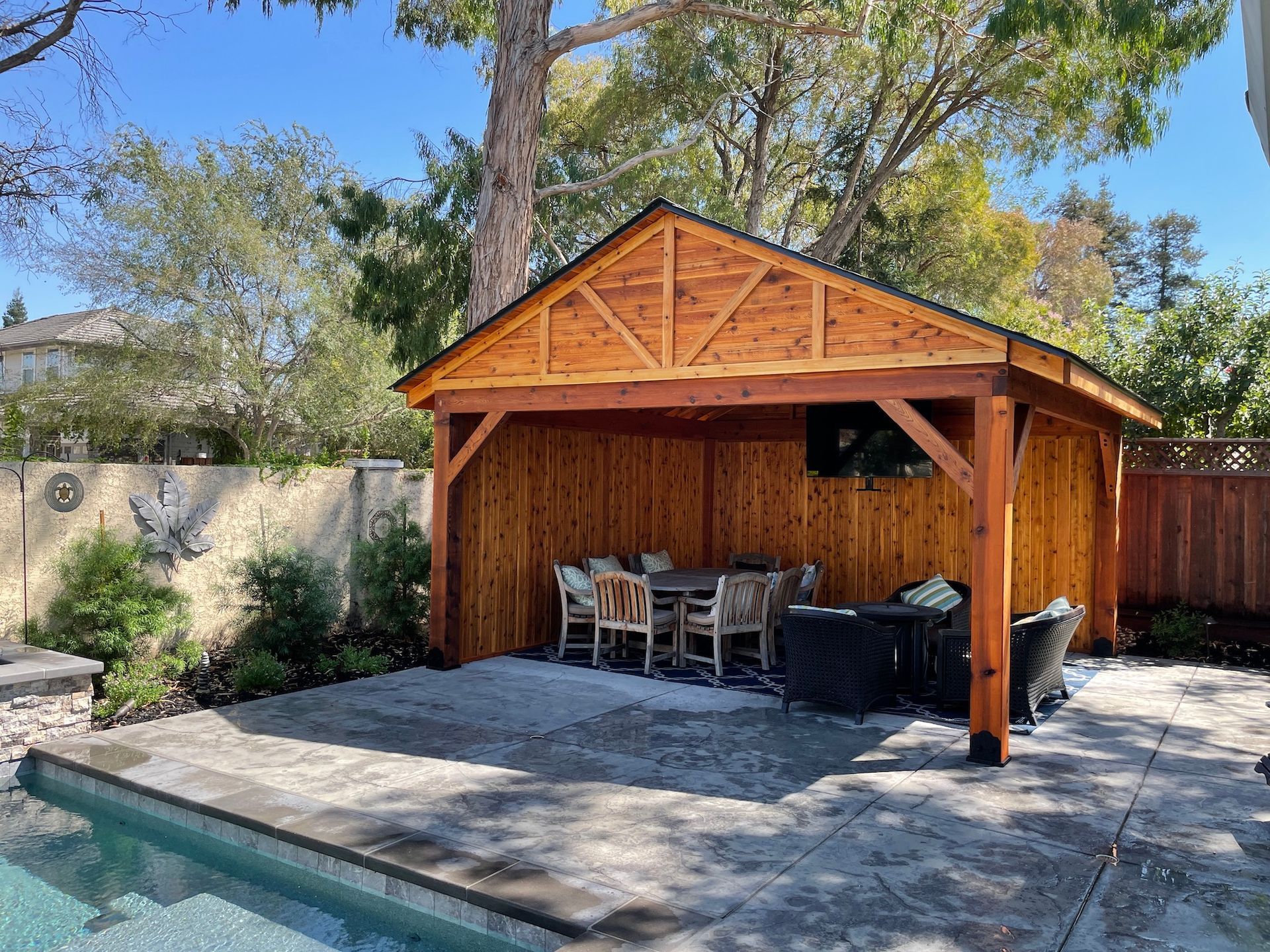 A wooden pavilion with a table and chairs in front of a pool.