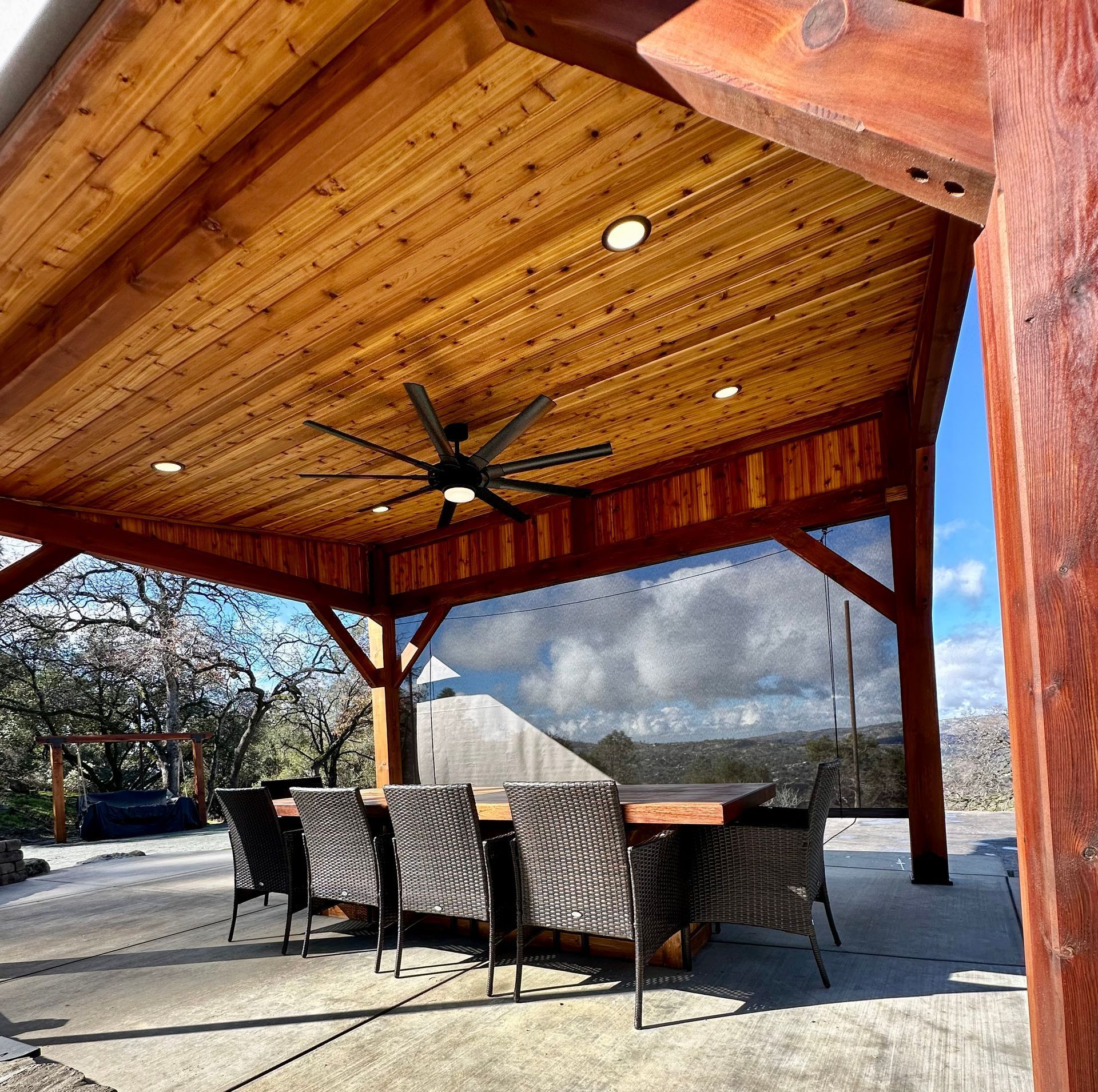 Wooden patio cover with dining set and ceiling fan, overlooking a scenic vista.