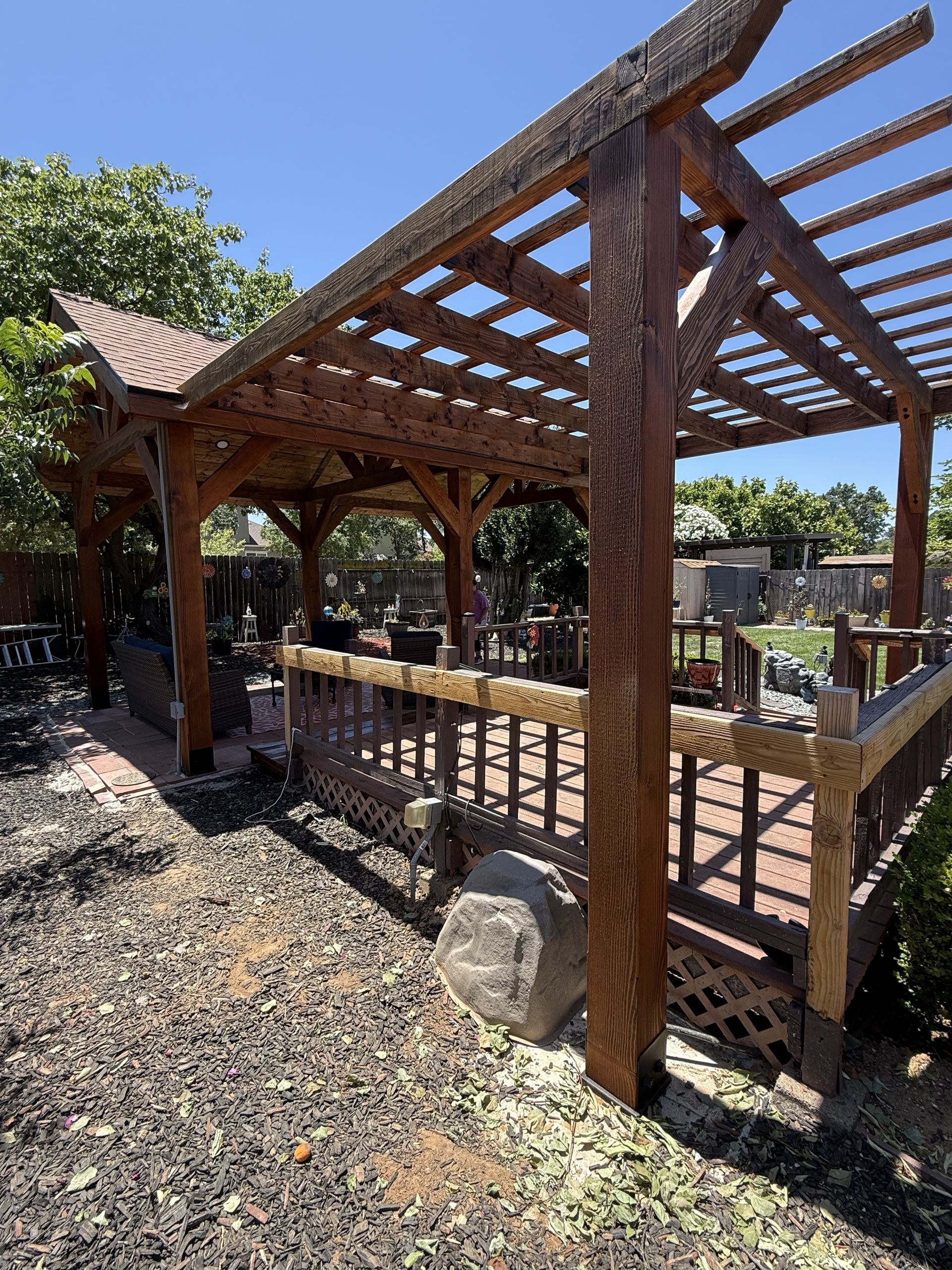 Wooden pergola with latticework and reddish-brown hues, in a backyard setting, under a blue sky.