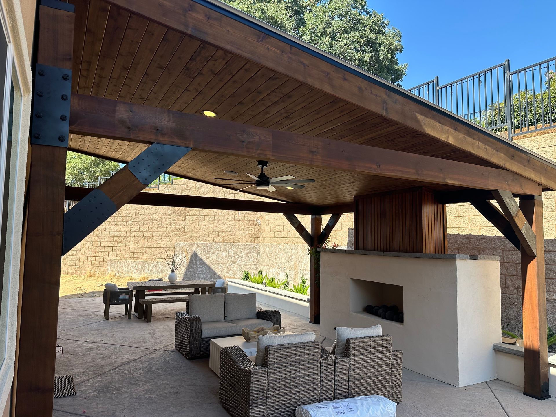 A patio with wicker furniture and a fireplace under a wooden pavilion.