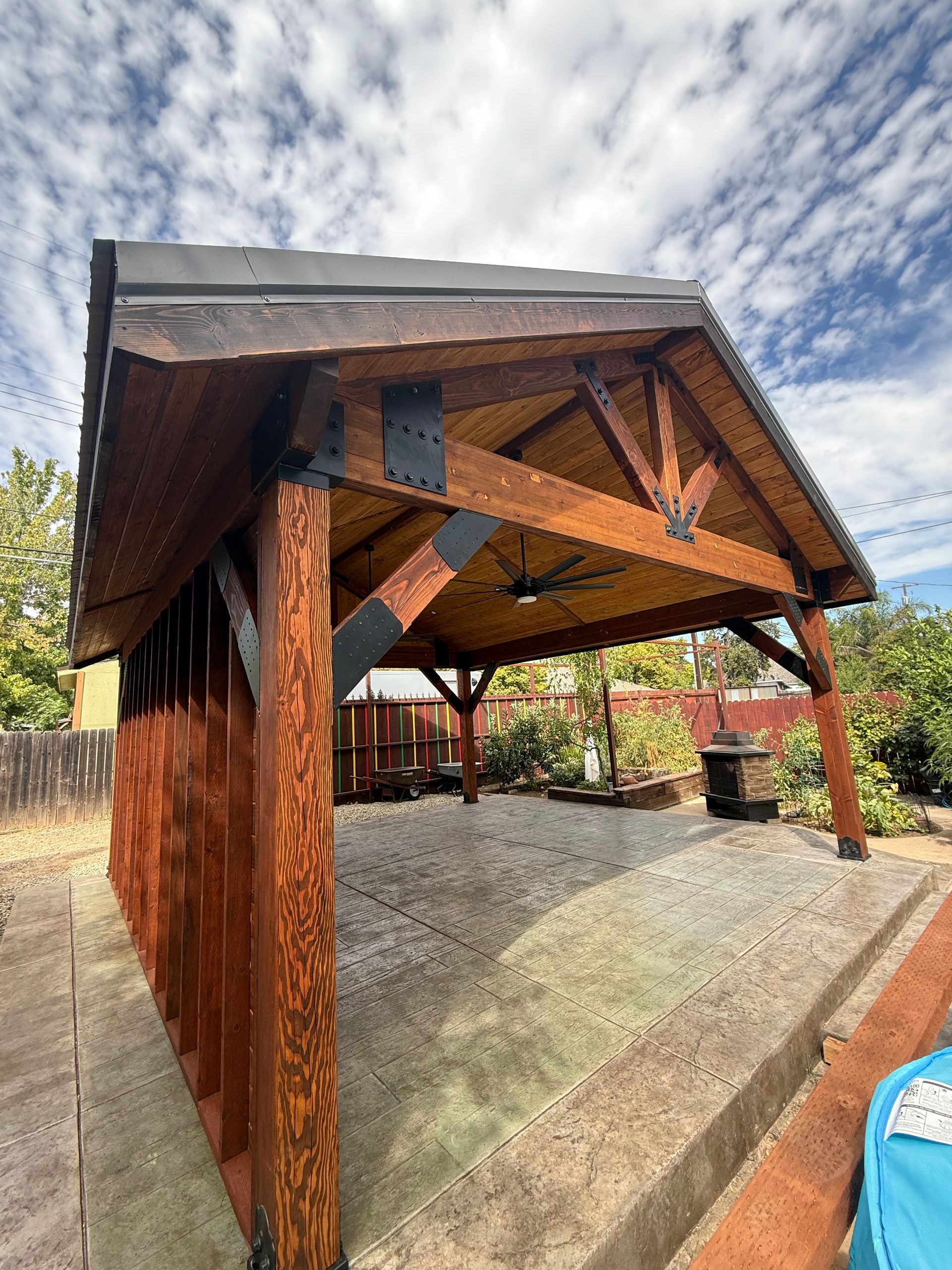 Wooden pergola with brown beams, a black roof, and gravel flooring in an outdoor setting.