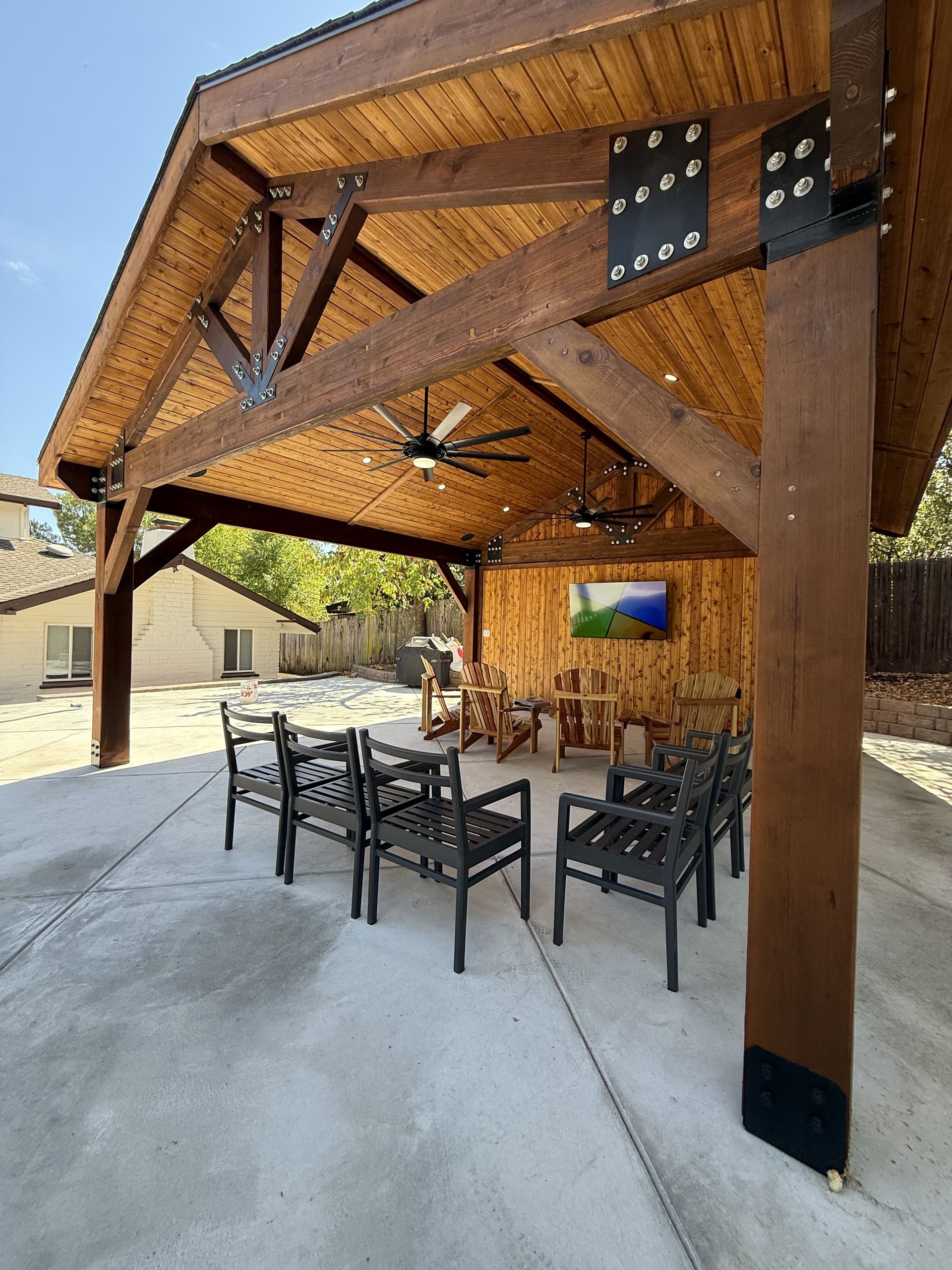 A covered outdoor patio with wooden beams, benches, and a television, set on a concrete surface.