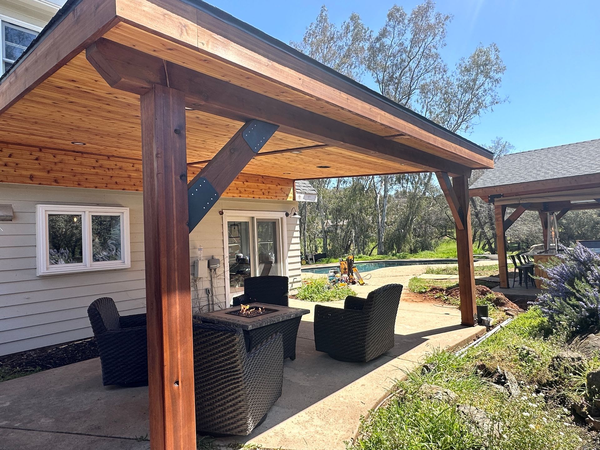A patio with a fire pit and chairs under a wooden roof.