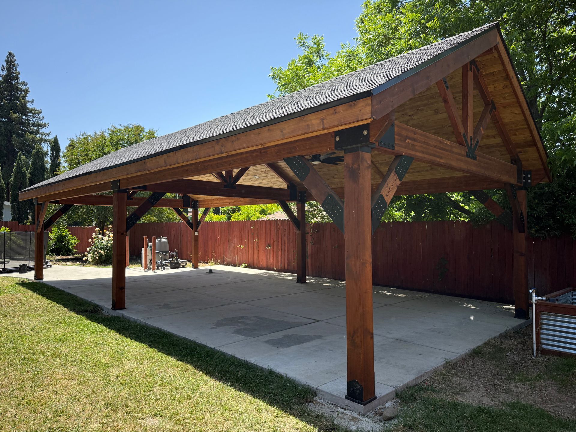 Wooden, brown-stained pavilion with metal roof in a backyard with a concrete patio.