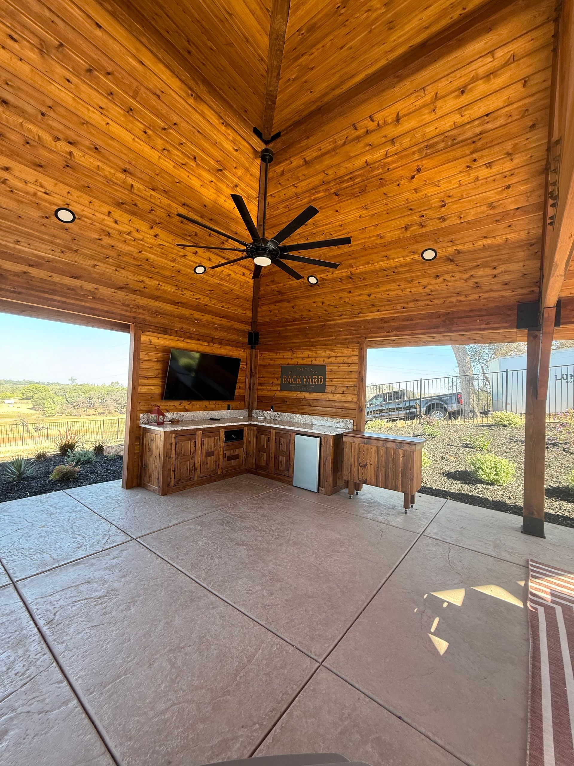 Outdoor kitchen under a wood-paneled roof with a ceiling fan, built-in bar and TV.