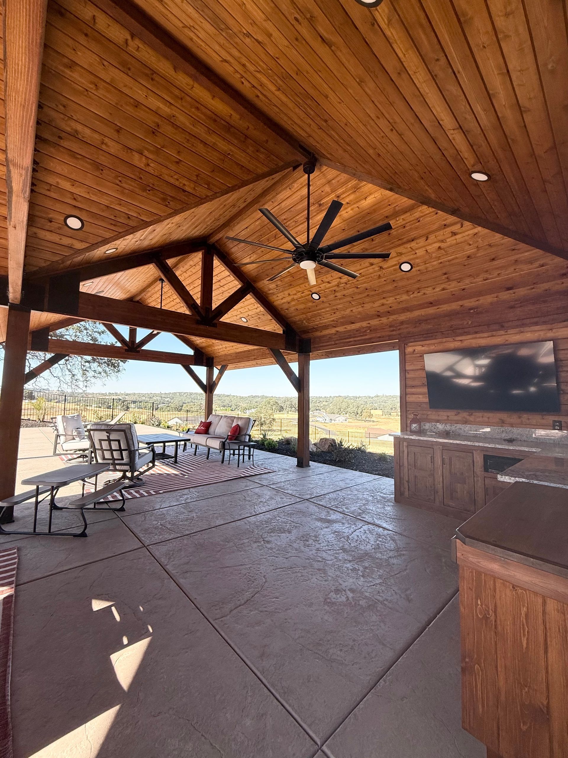 Outdoor patio with wooden beams, fan, seating, a TV, and a view.