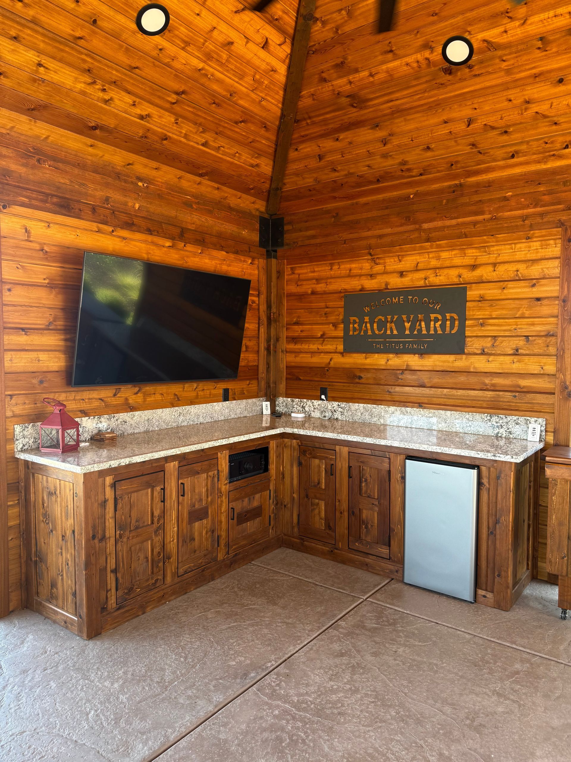 Outdoor kitchen with wood cabinetry, stone countertops, TV, and mini-fridge under a wood-paneled ceiling.