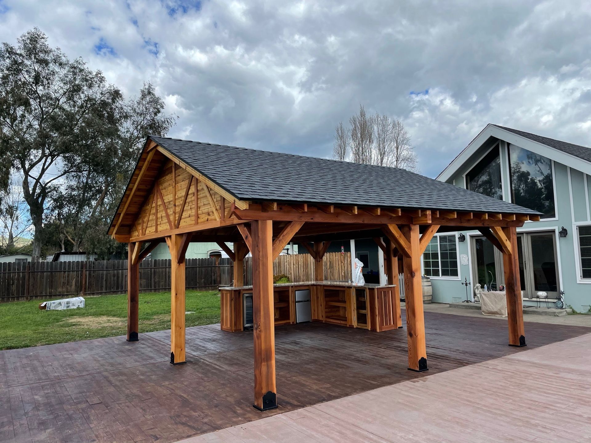 A wooden pavilion with a slate roof is in front of a house.