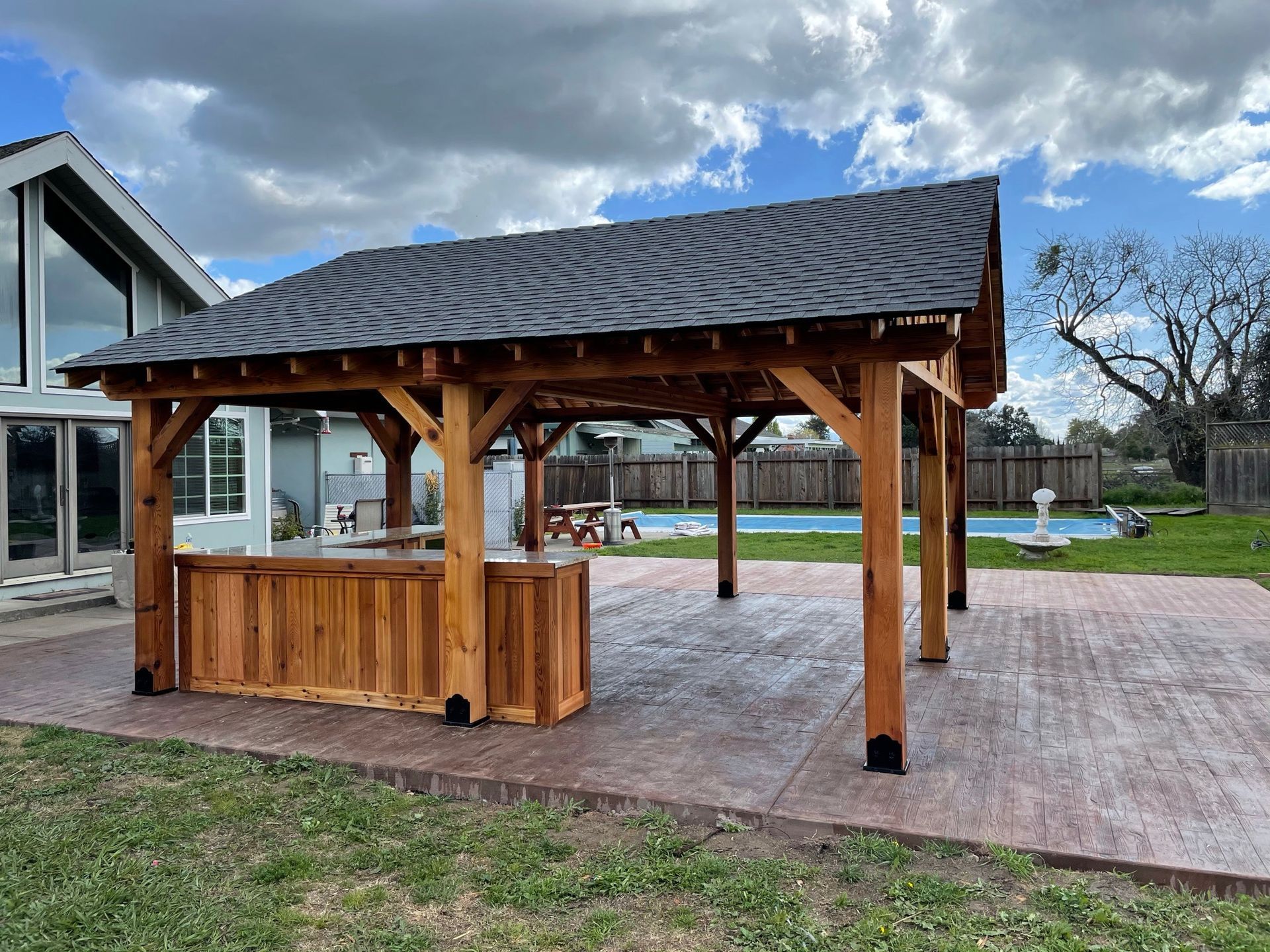 A wooden gazebo with a bar underneath it is in the backyard of a house.