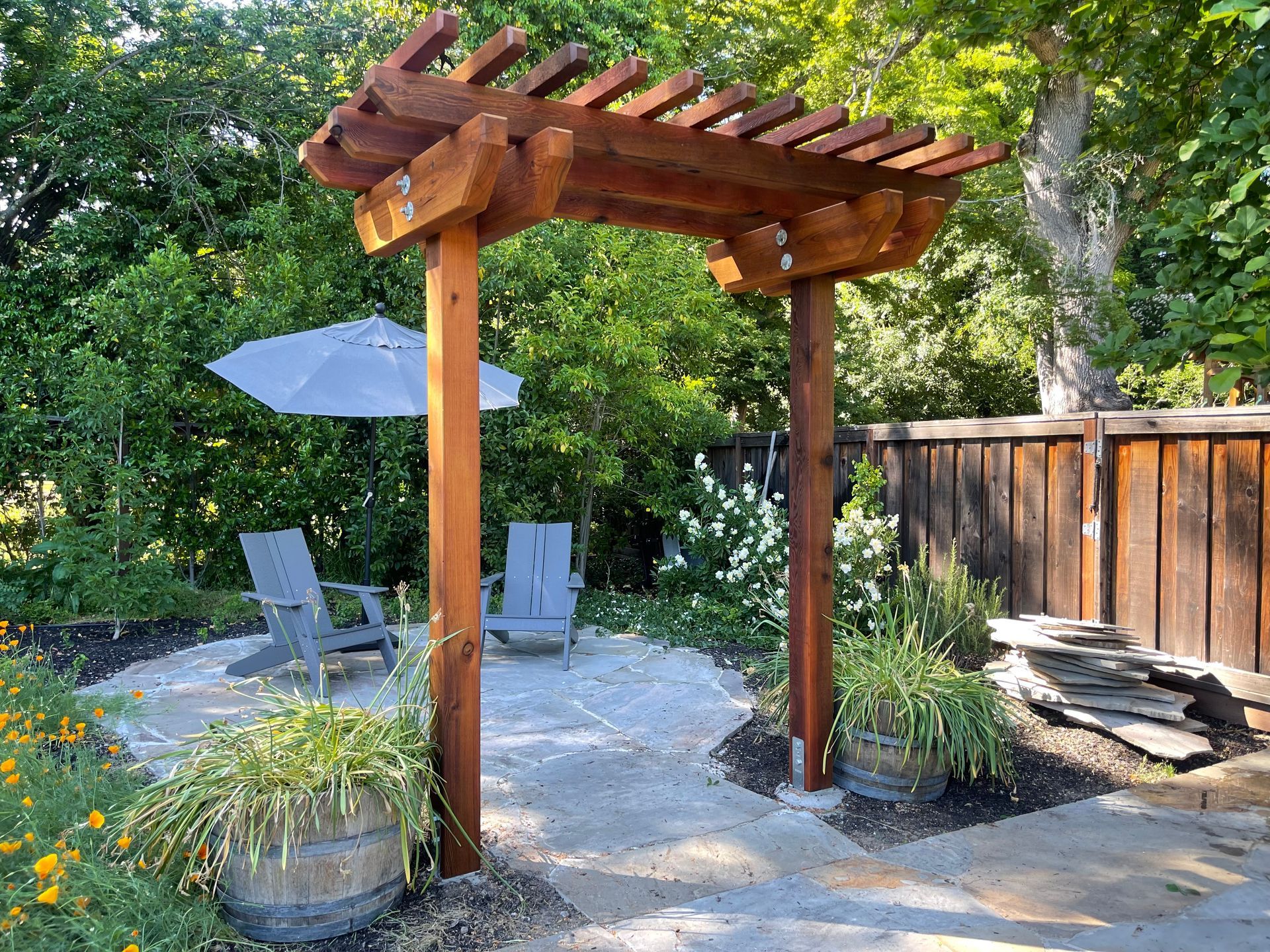 A wooden pergola with chairs and an umbrella in a backyard.