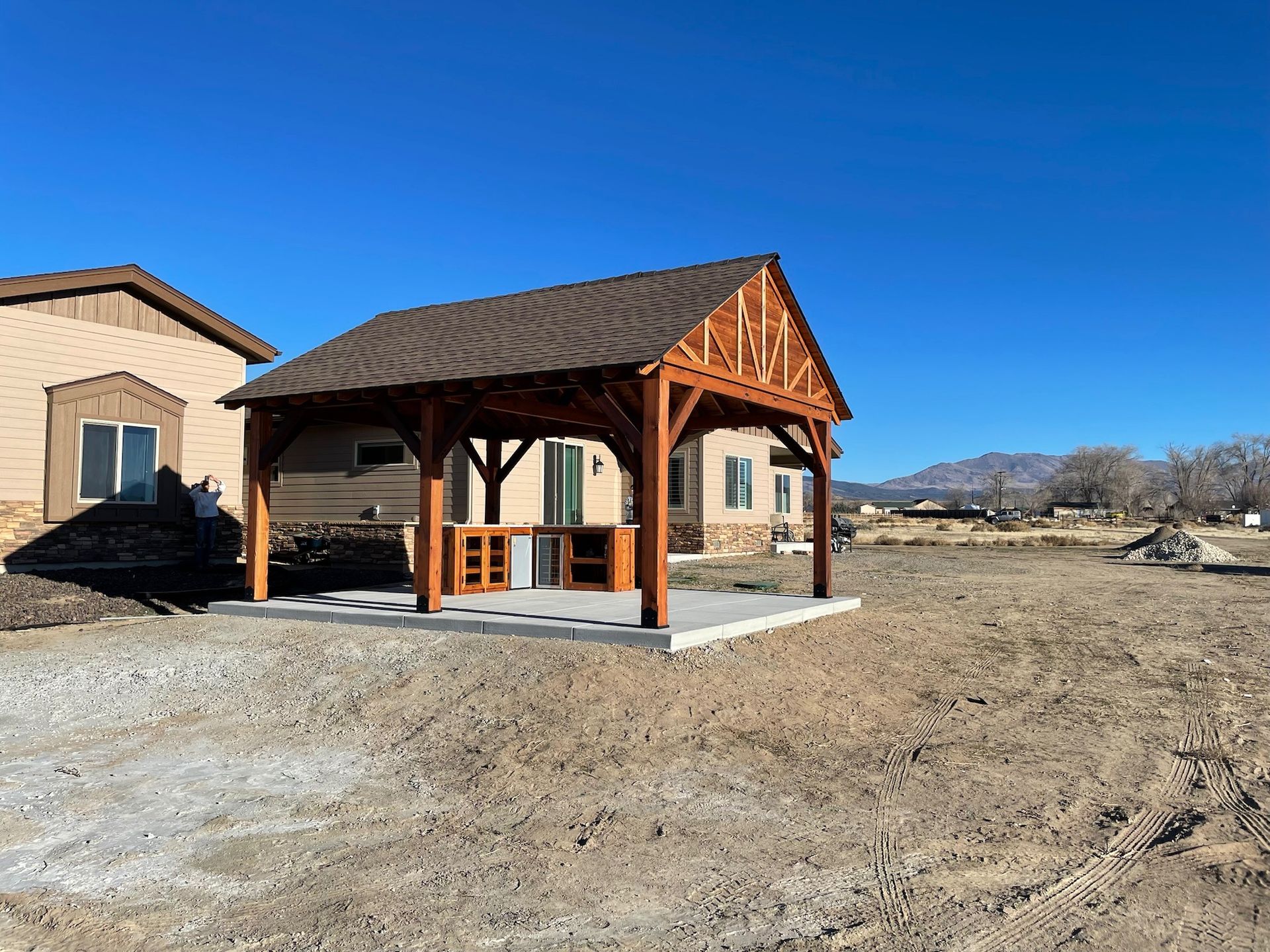 A wooden gazebo is sitting in the middle of a dirt field in front of a house.