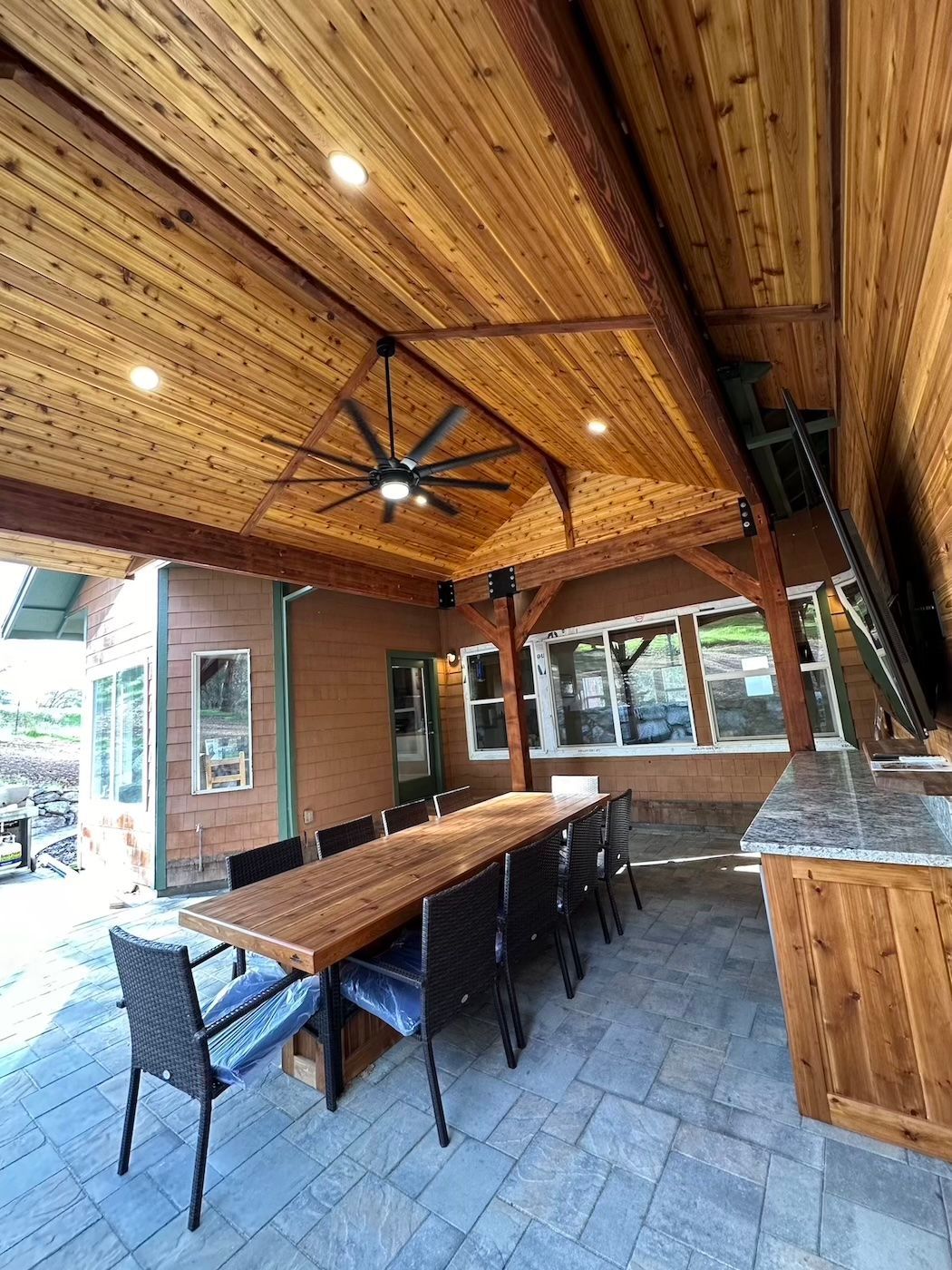 A large wooden table and chairs under a wooden ceiling with a ceiling fan.