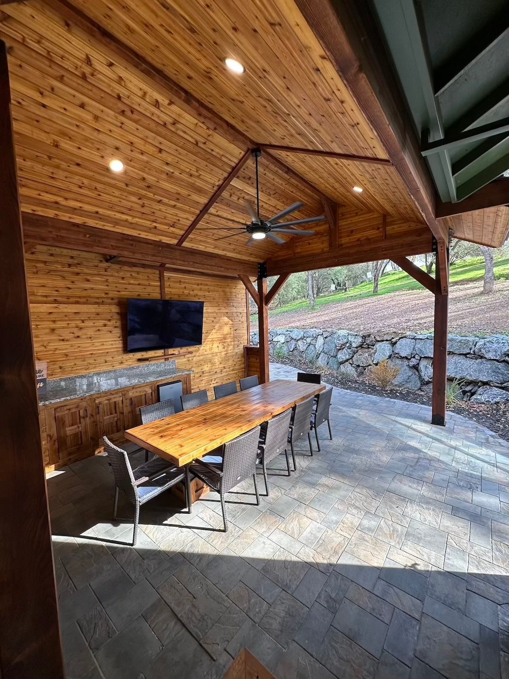 A large wooden table and chairs under a wooden roof with a flat screen tv mounted on the wall.