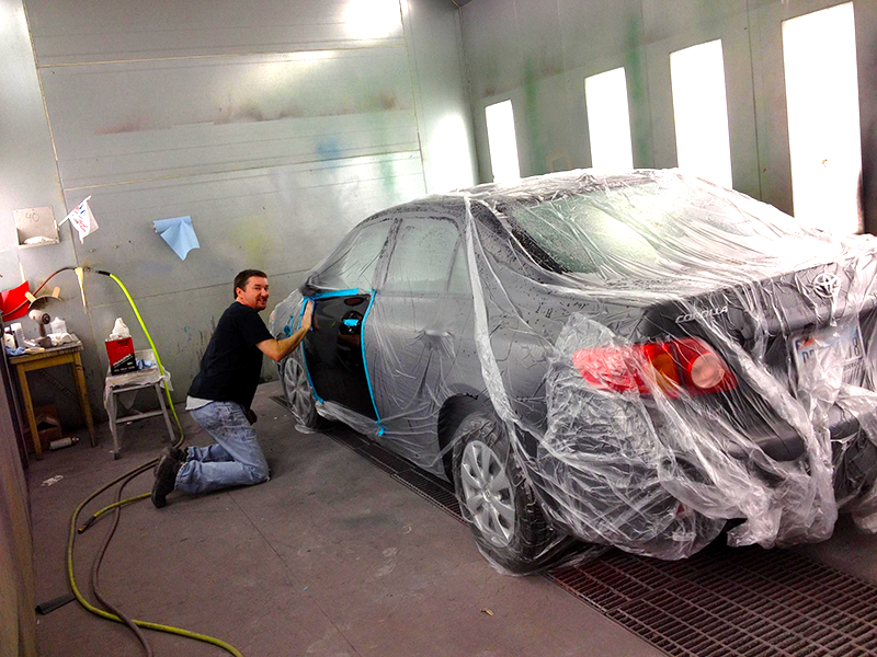 A man is kneeling next to a car wrapped in plastic in a paint booth.