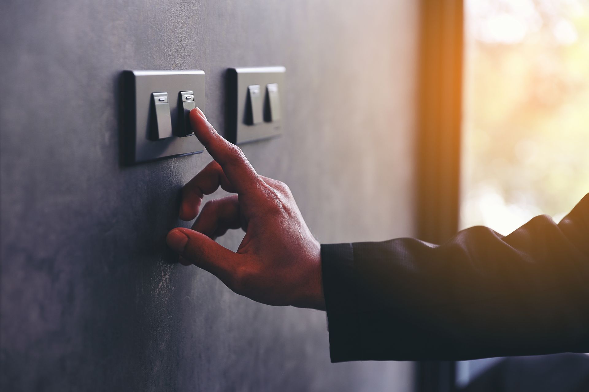 Hand pressing a light switch on a wall panel, with warm sunlight in the background