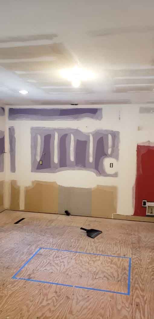 Room under construction, exposed drywall with tape and plaster. Wood floor with blue tape outline.