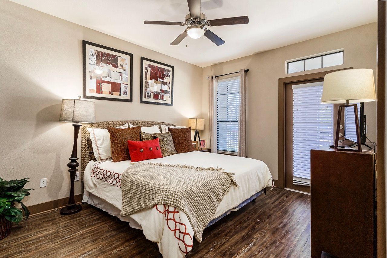 Bedroom with a king bed, multiple throw pillows, two lamps, ceiling fan, dresser, and windows with blinds.