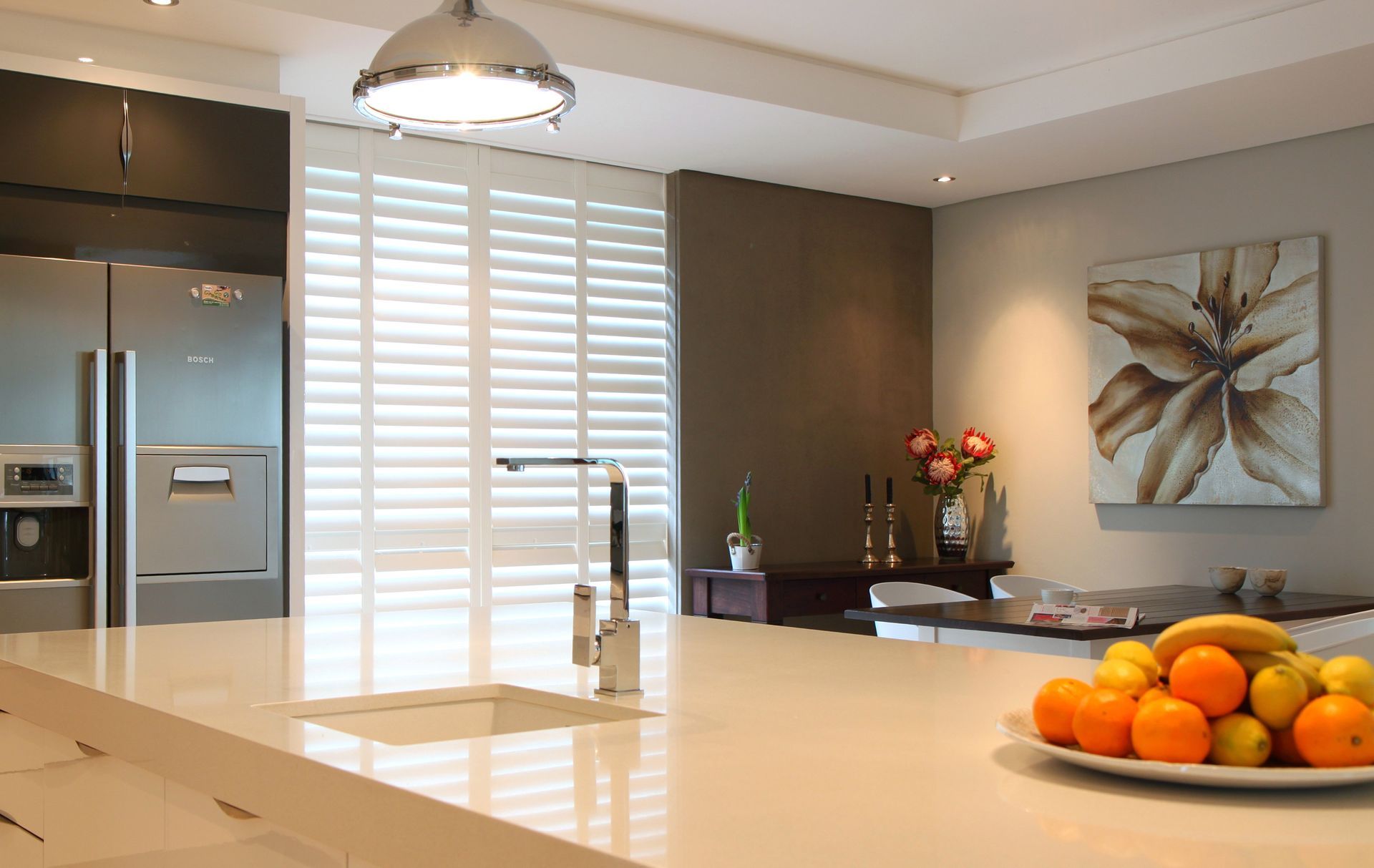 Modern kitchen with a white island, stainless steel appliances, and fruit bowl. White shutters cover the window.