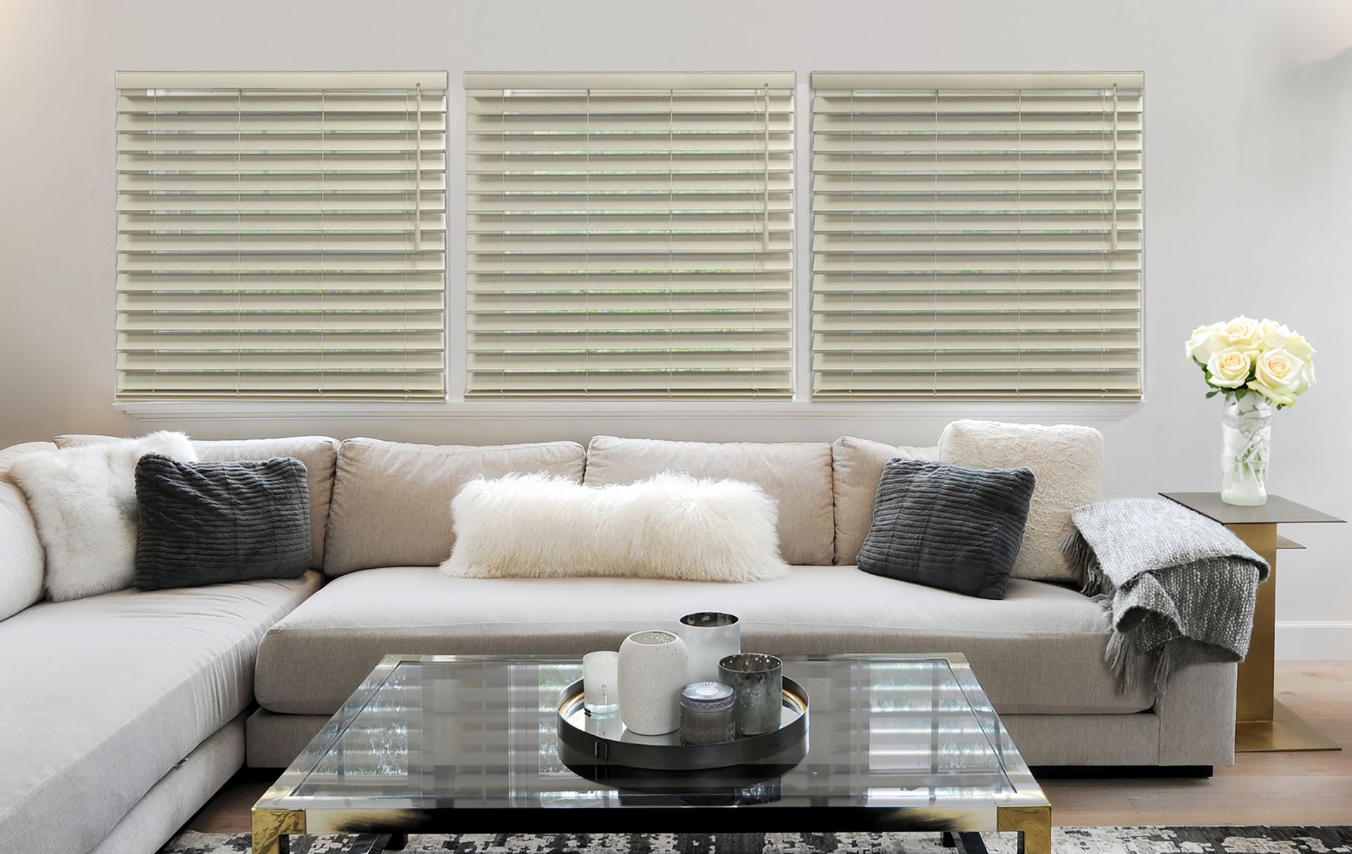 Living room with light-colored blinds, sectional sofa with pillows, glass coffee table, and a side table with flowers.
