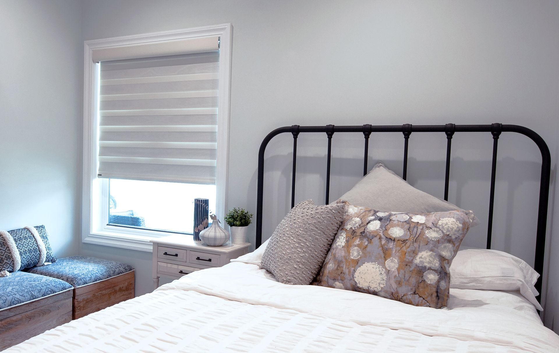 Bedroom with bed, pillows, window, and small bedside table; pale gray walls and neutral tones.