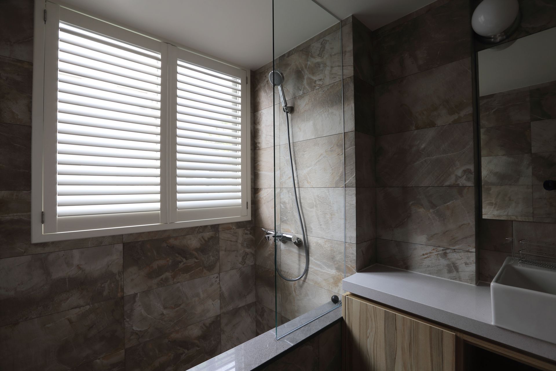 Bathroom with marble-like tile, shower, window with shutters, and vanity with a sink.
