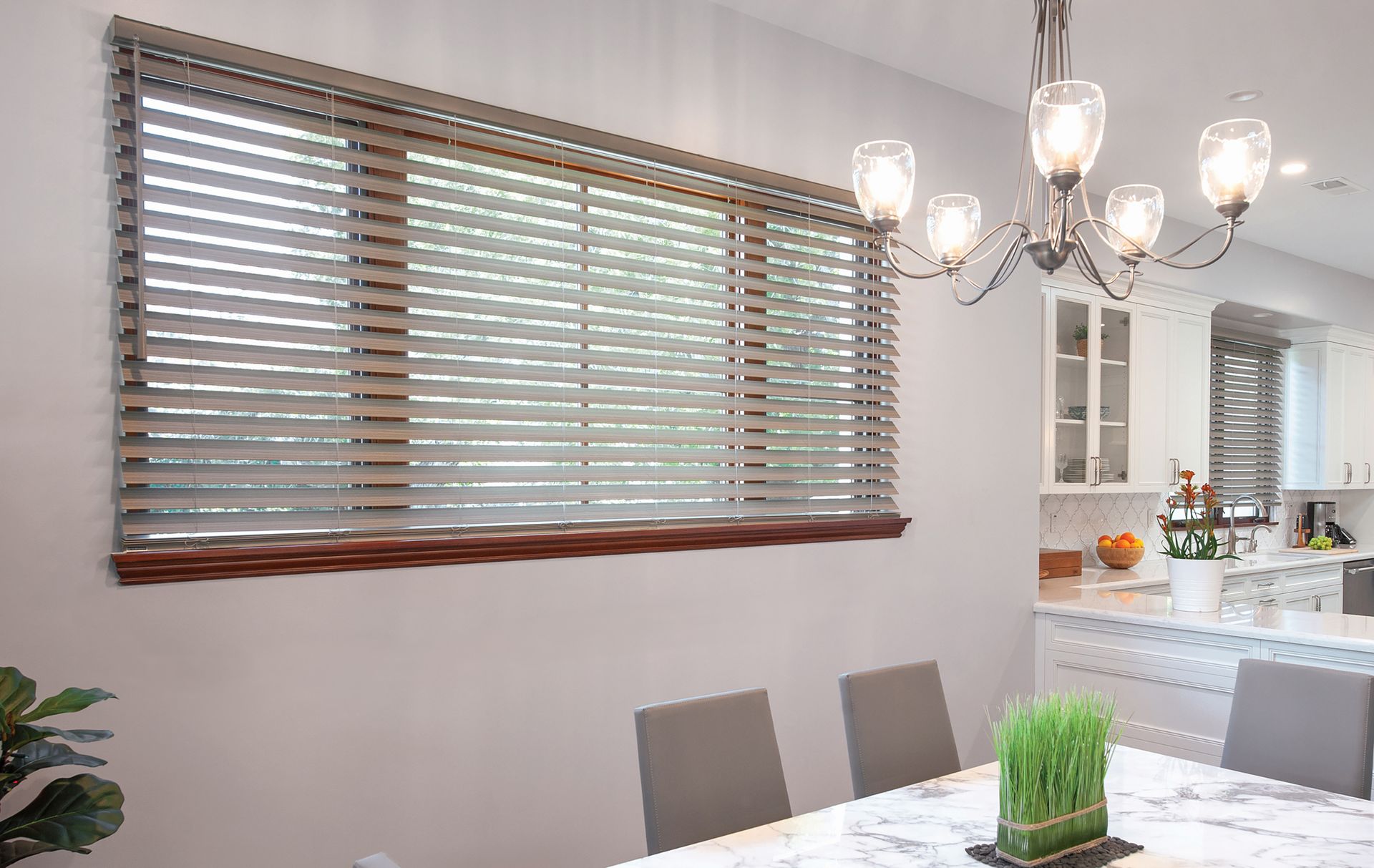 Dining room with gray blinds on a large window, chandelier above a marble table.
