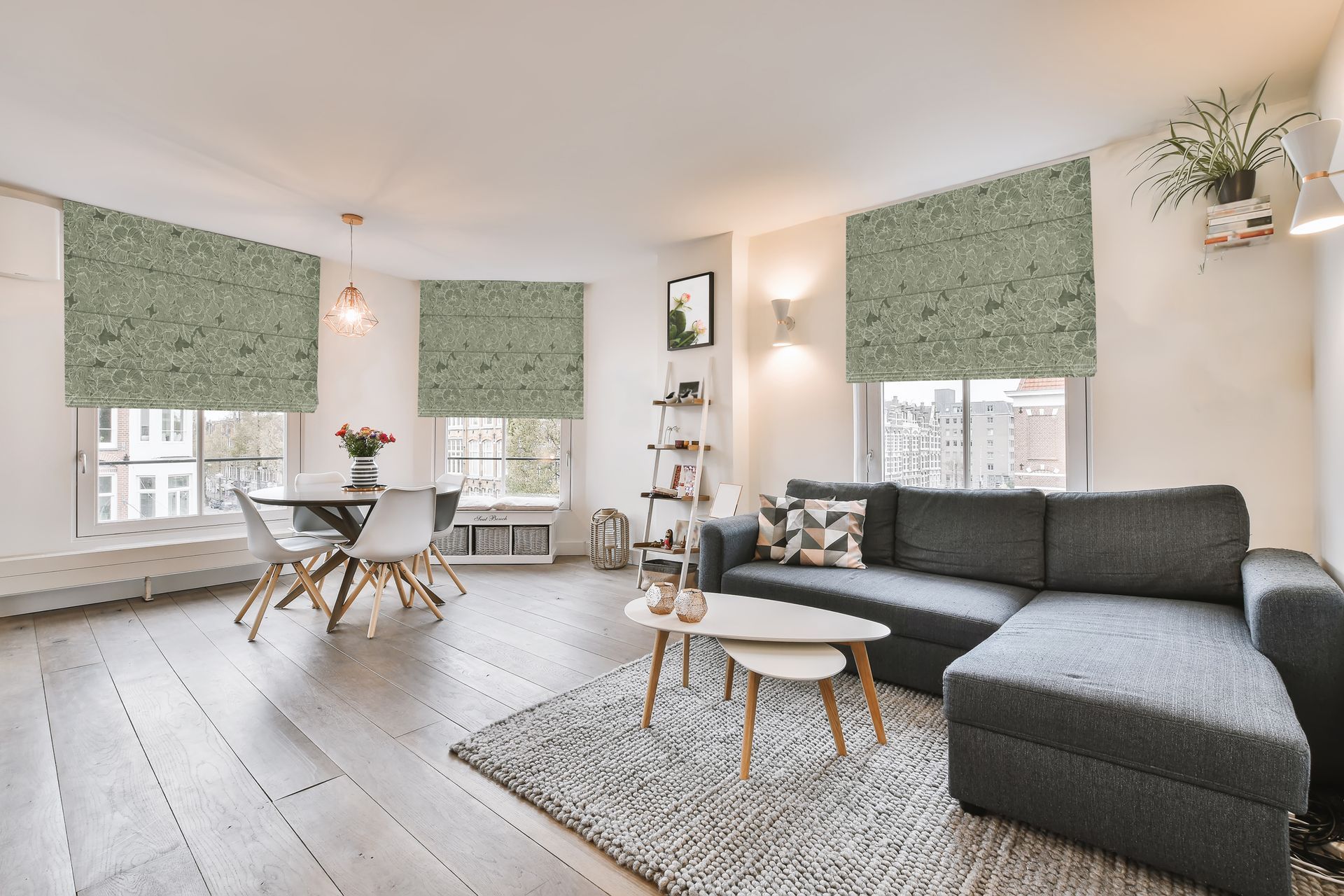 Living room with dark gray sectional sofa, round dining table, patterned window shades, and area rug.
