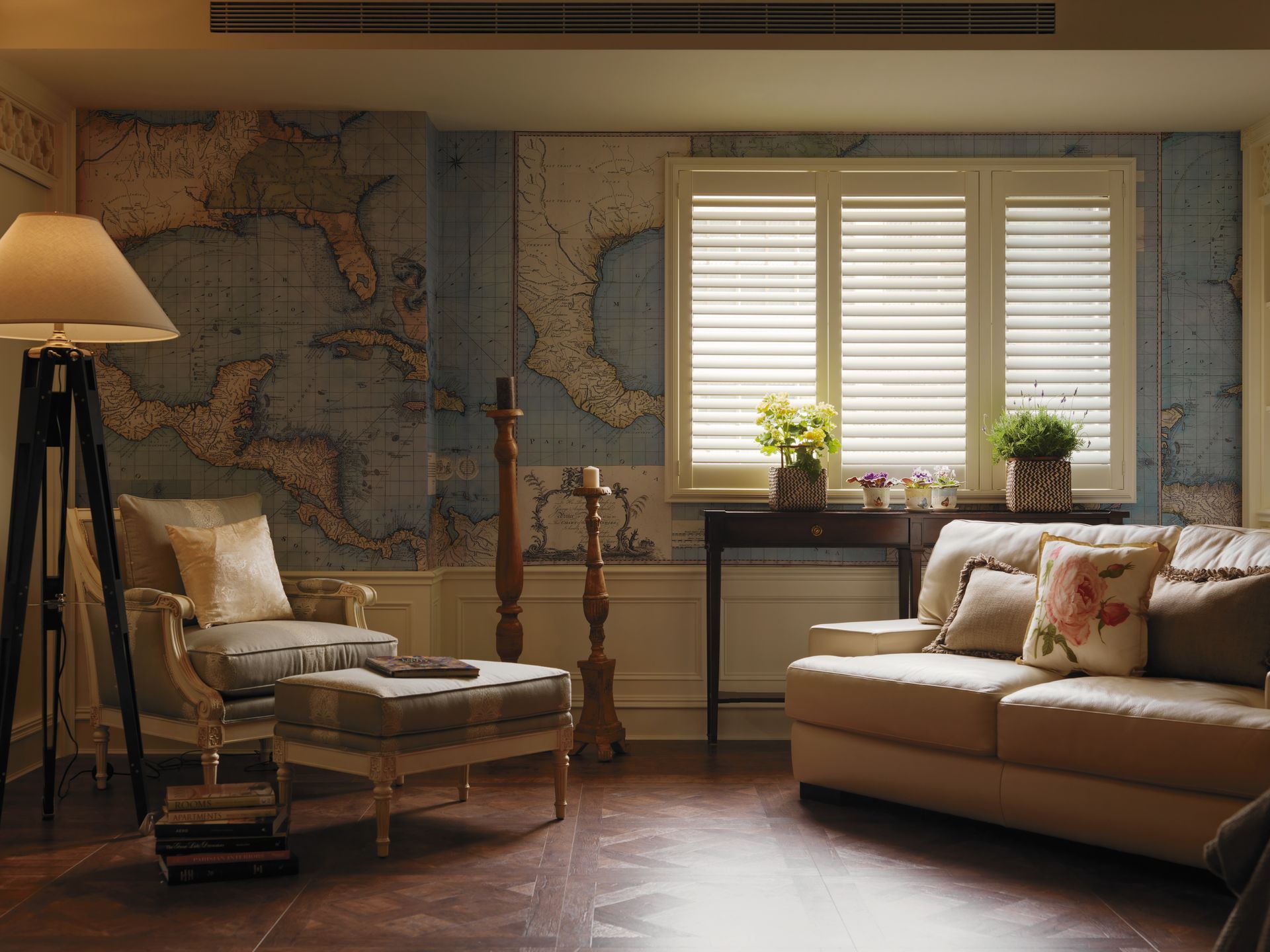 Living room with map-patterned wall, beige couch, armchair, ottoman, and large window with white shutters.