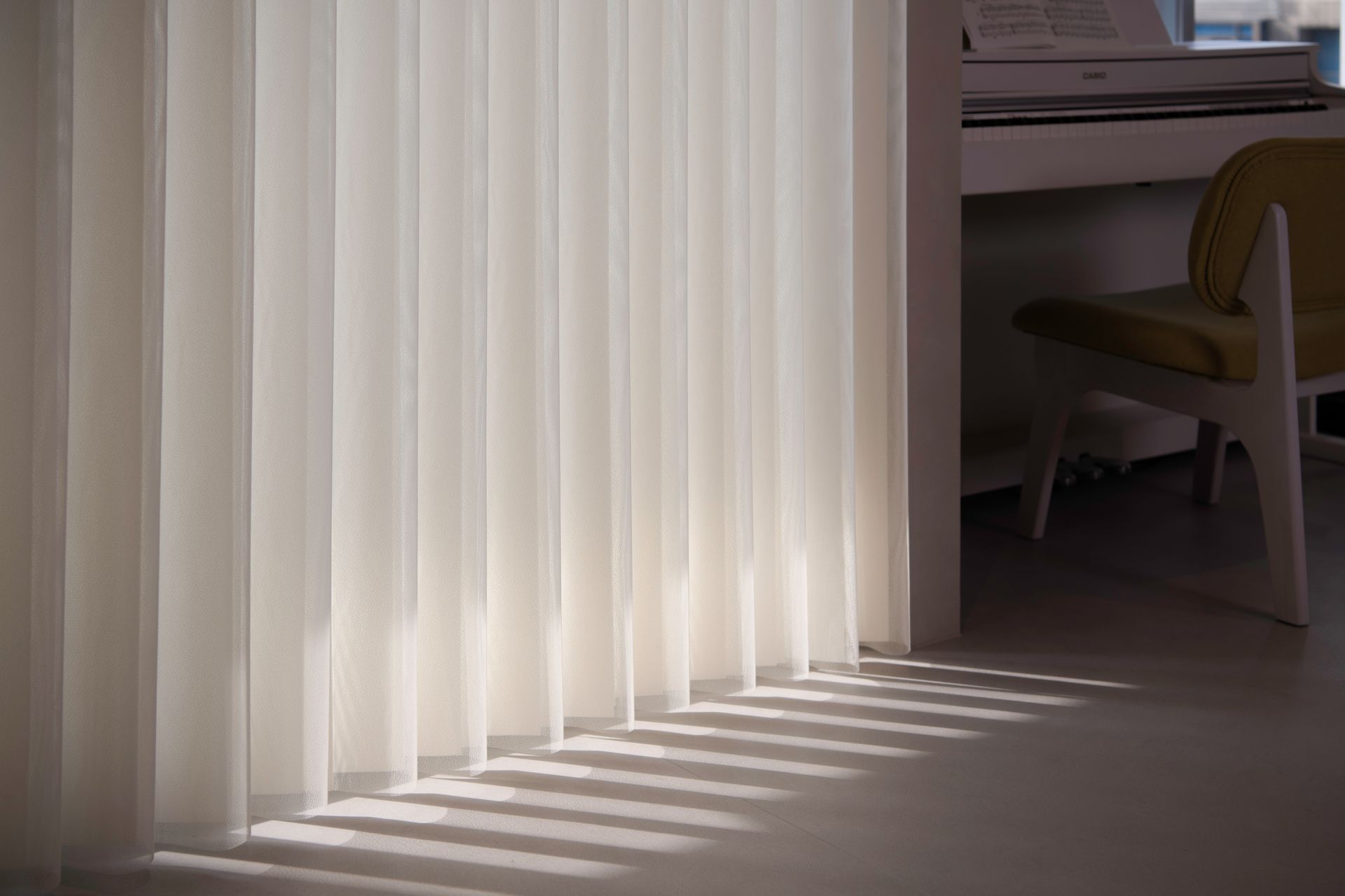 Vertical blinds casting shadows on a light-colored floor, with a piano and chair in the background.