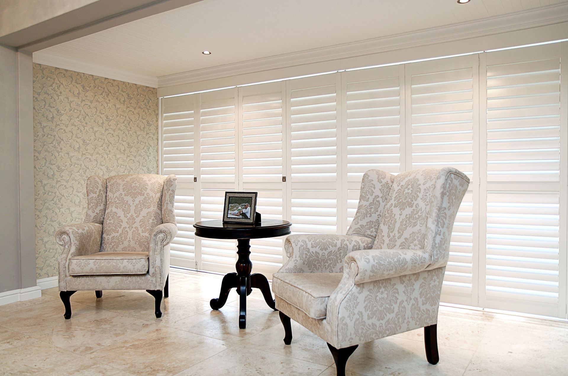 Two patterned armchairs face each other near a round table; white shutters in the background.