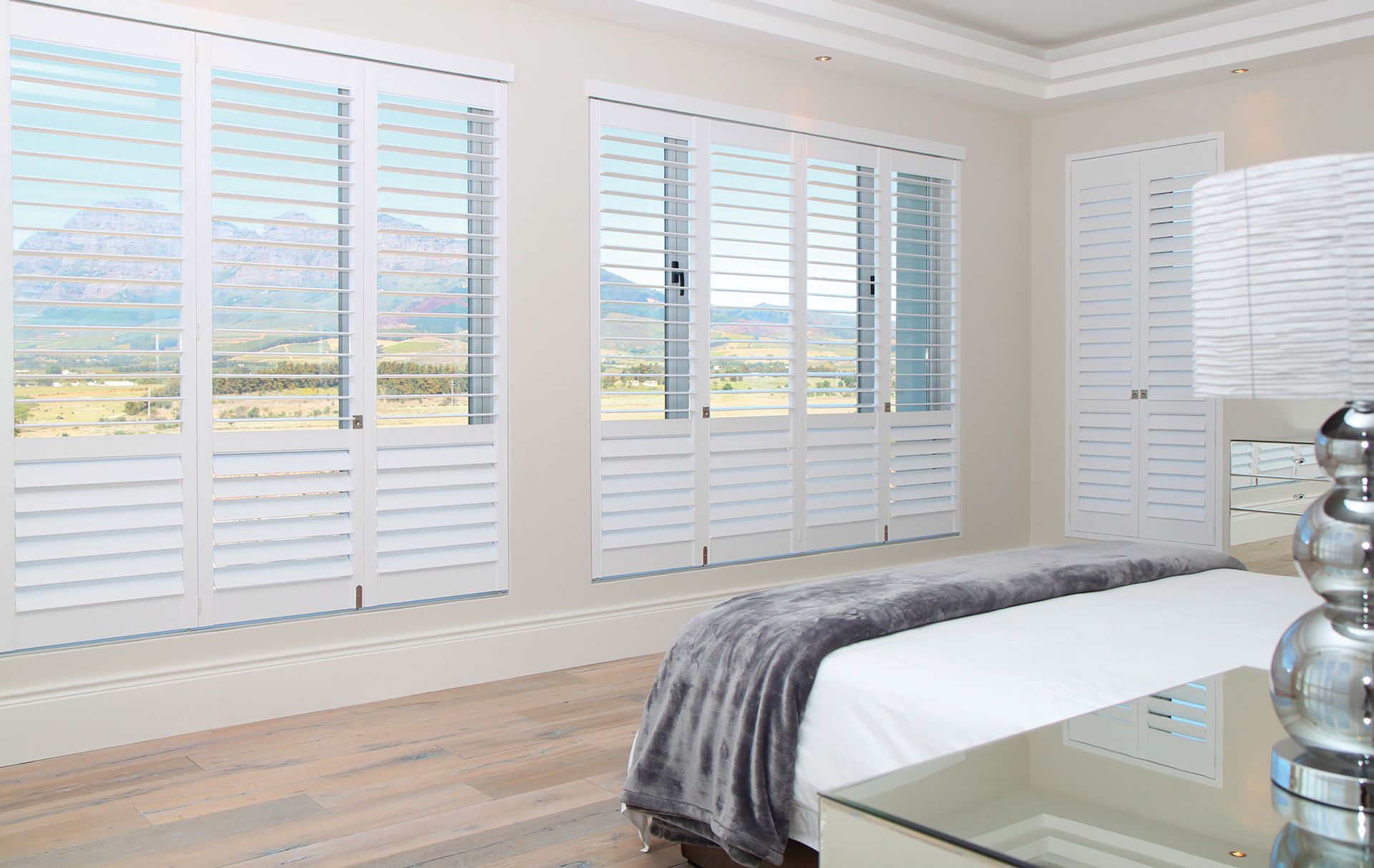 Bedroom with white shutters, bed with gray blanket, light wooden floors, and view of mountains.
