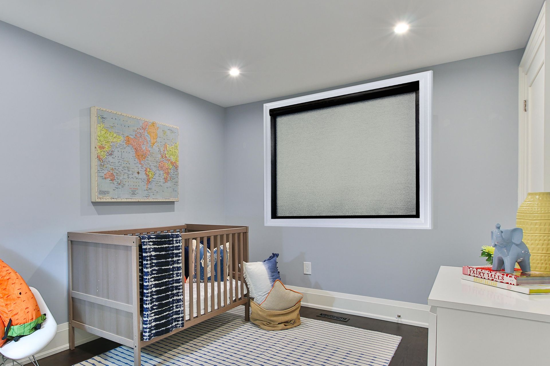 Baby's nursery with crib, world map art, window with shade, and a white dresser. The walls are light blue.