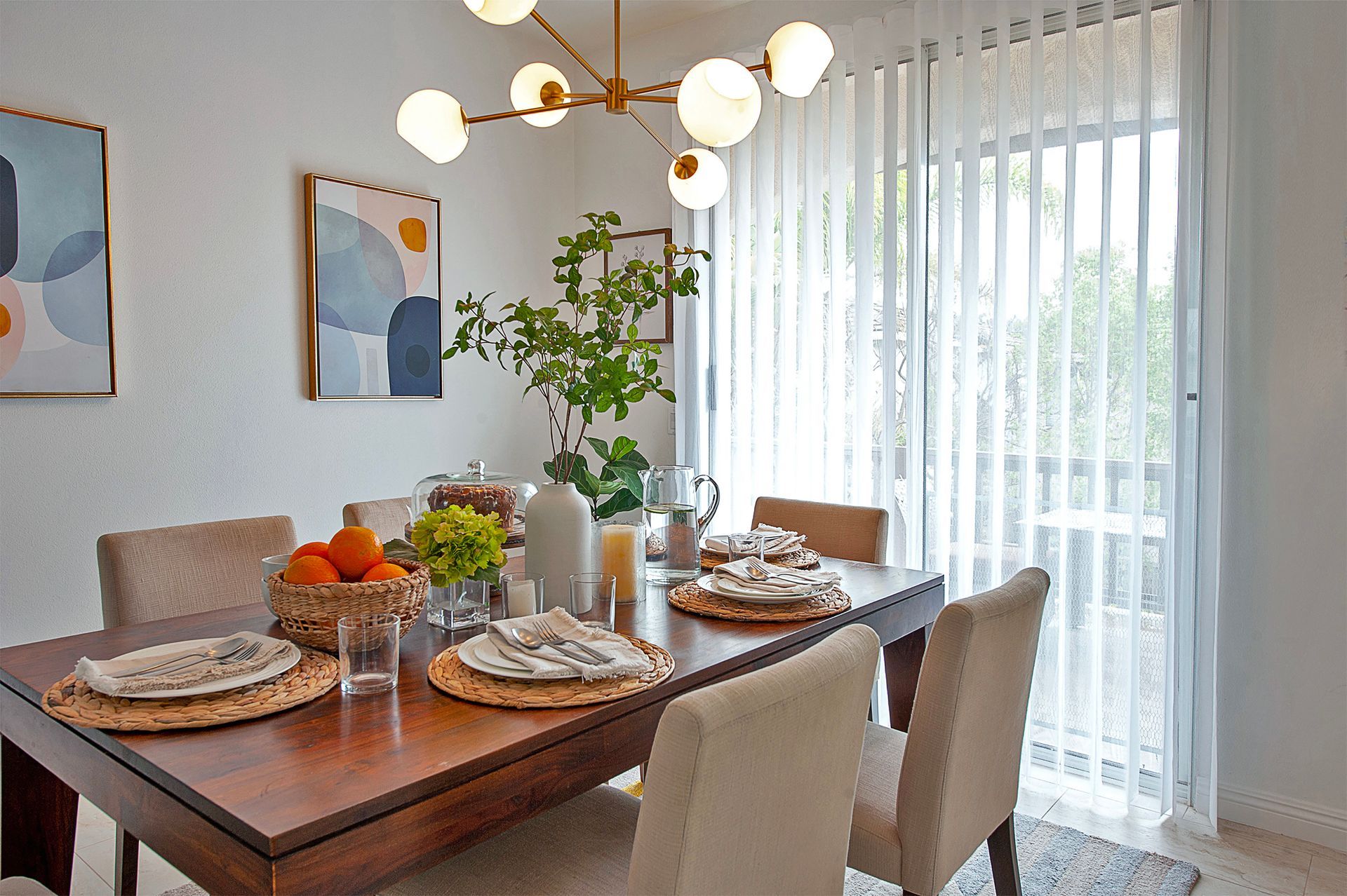 Dining room with a wooden table set for a meal. Soft lighting, art, and a vase of greenery.